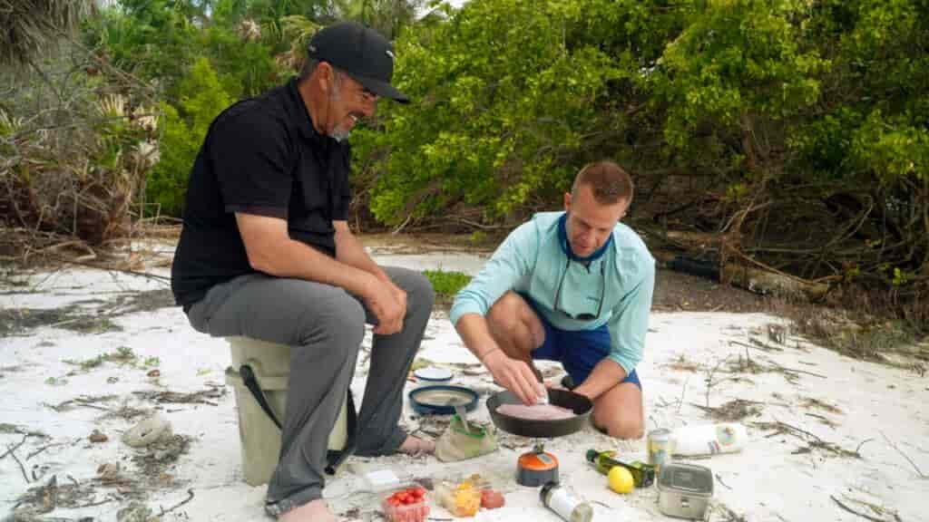 Chef Rob and Chef Corso frying fish in a pan on a camp stove on the beach.